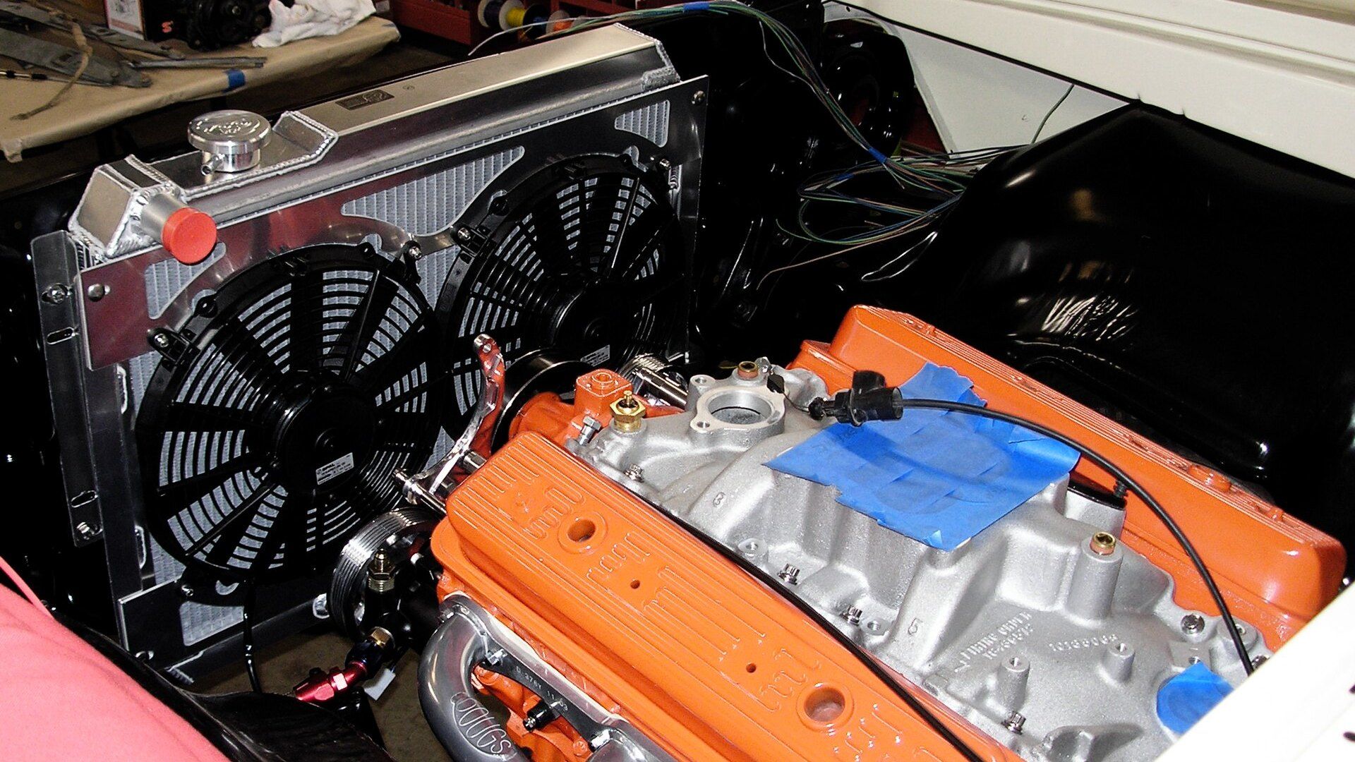 A full aluminum radiator with welded aluminum tanks and a polished filler cap, fitted with two black electric cooling fans in front of a V8 engine with orange valve covers
